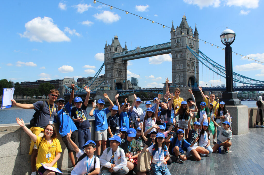 Discovery Summer students waving and wearing blue caps on a London excursion to Tower Bridge
